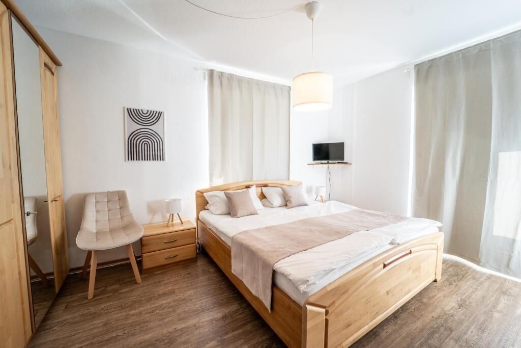 A modern bedroom with a wooden bed, a bedside table, a chair, a TV mounted on the wall, and a window with curtains.