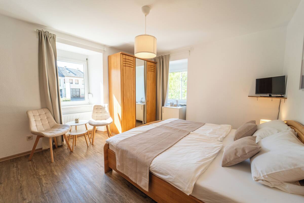 Une chambre à coucher avec un lit en bois, une chaise, une petite table, un téléviseur et une fenêtre avec des rideaux.