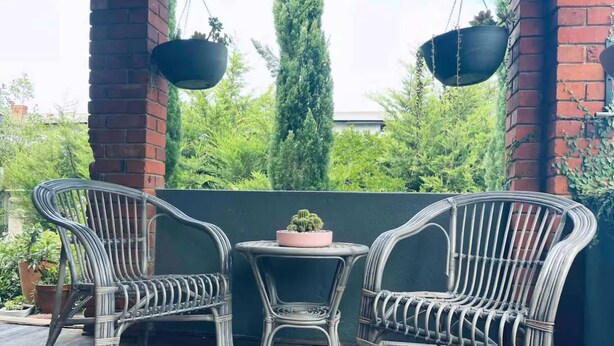 A balcony with wicker chairs and a small table, surrounded by greenery and a brick wall.
