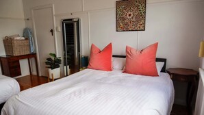 A bedroom with a large bed, two pillows, a nightstand, a mirror, a door, a plant, and a wall decoration.