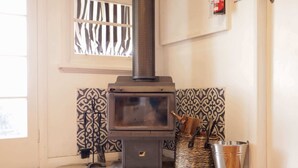 A wood-burning stove in a corner with patterned tiles, a metal bucket, and a window with blinds.