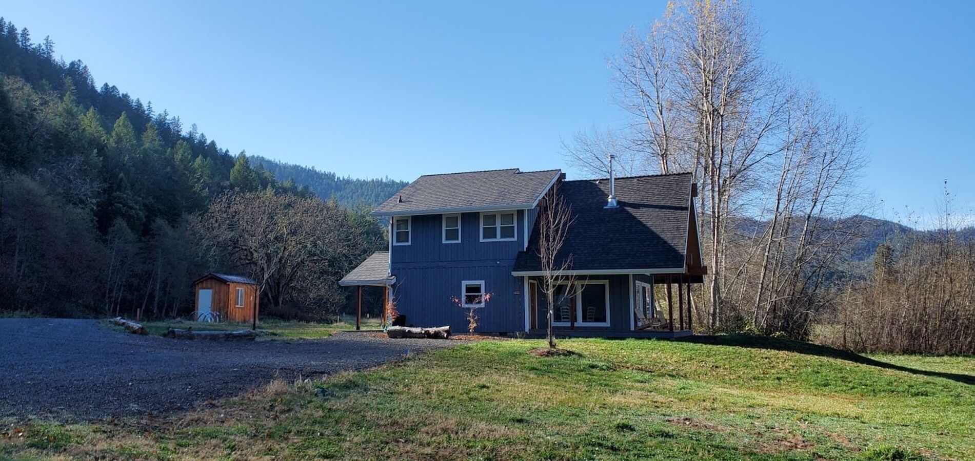 A two-story house with a dark blue exterior, a small wooden shed, and a clear view of a forested hillside in the background.