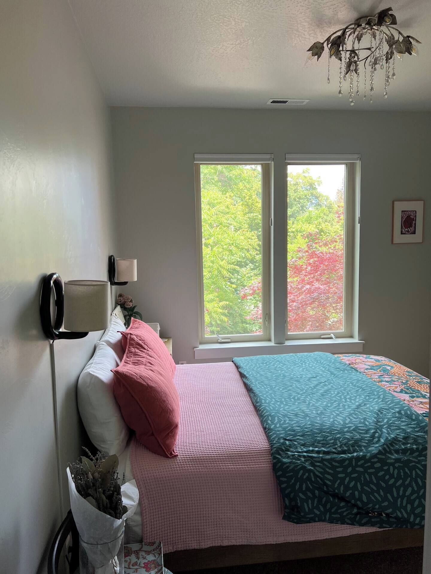 A bedroom with a bed, a window with a view of trees, a lamp, and a decorative chandelier.