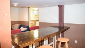 Een kamer met een houten eettafel, twee krukken, een rode stoel en een bank met kussens.
