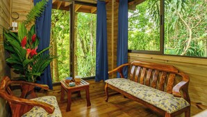 A wooden room with a bench, two chairs, a small table, and a large window overlooking greenery. - Peaceful Jungle Cabin at Ginger Retreat with River Sounds & Fast Internet (Quepos)