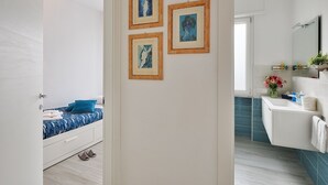 Un bagno moderno con un mobile vanitario bianco, uno specchio e un lavandino. Alle pareti sono presenti opere d'arte incorniciate e, nella stanza accanto, un letto con un cuscino blu.
