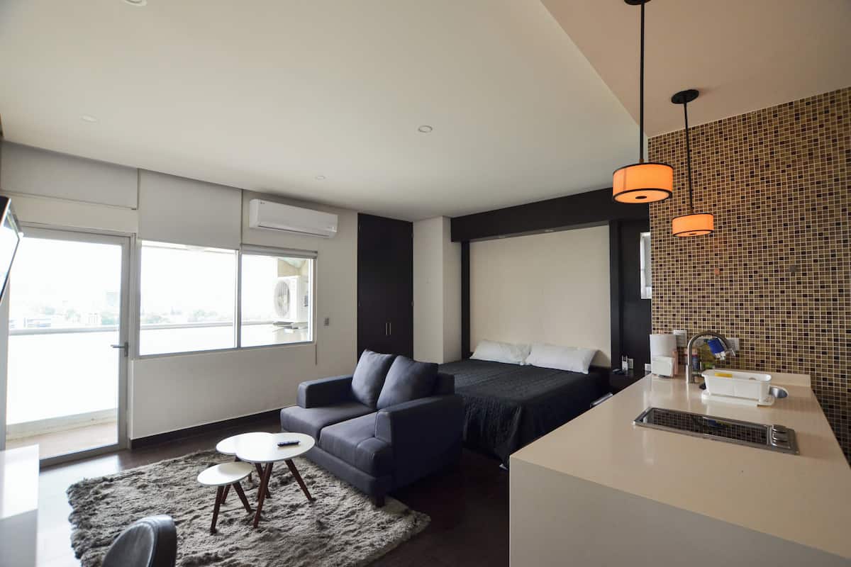 A modern hotel room with a sofa, a small table, a bed, and a kitchen area.