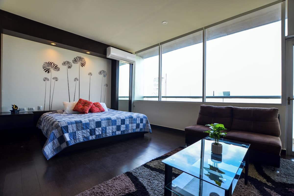 A modern bedroom with a large bed, a sofa, a glass coffee table, and a view of the city through large windows.