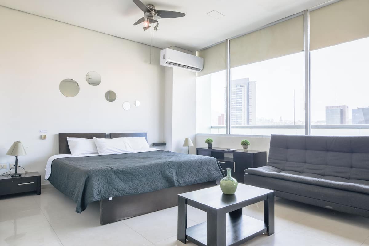 A modern hotel room with a bed, a sofa, a table, and a large window offering a city view.