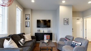 Un salon comprenant un canapé, des fauteuils, une table basse, un téléviseur à écran plat et un meuble de rangement noir pour les appareils électroniques.
