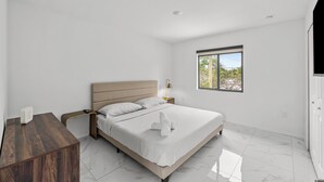 A bedroom with a bed, bedside table, lamp, and a window with a view. - Urban Sabor - Modern Miami - King Beds - Fenced Yard - Parking (Miami)