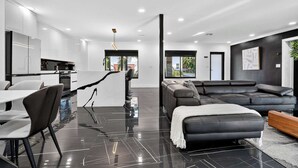 A modern living space with a kitchen, dining area, and a large black leather sofa. - Coral Lane by Rove Travel Heated Pool & BBQ (Fort Lauderdale)
