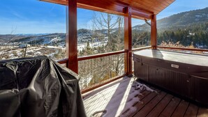 A wooden deck with a hot tub and a covered grill area, overlooking a snowy landscape with trees and hills. - Cozy Rustic Escape (Park City)