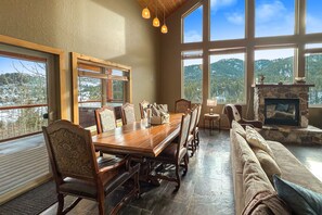 A spacious dining area with a long wooden table and chairs, a stone fireplace, and large windows offering mountain views. - Cozy Rustic Escape (Park City)