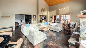 A spacious living room with a high ceiling, stone flooring, and a fireplace. There are multiple seating areas with sofas and chairs, and a dining table with chairs. - Cozy Rustic Escape (Park City)