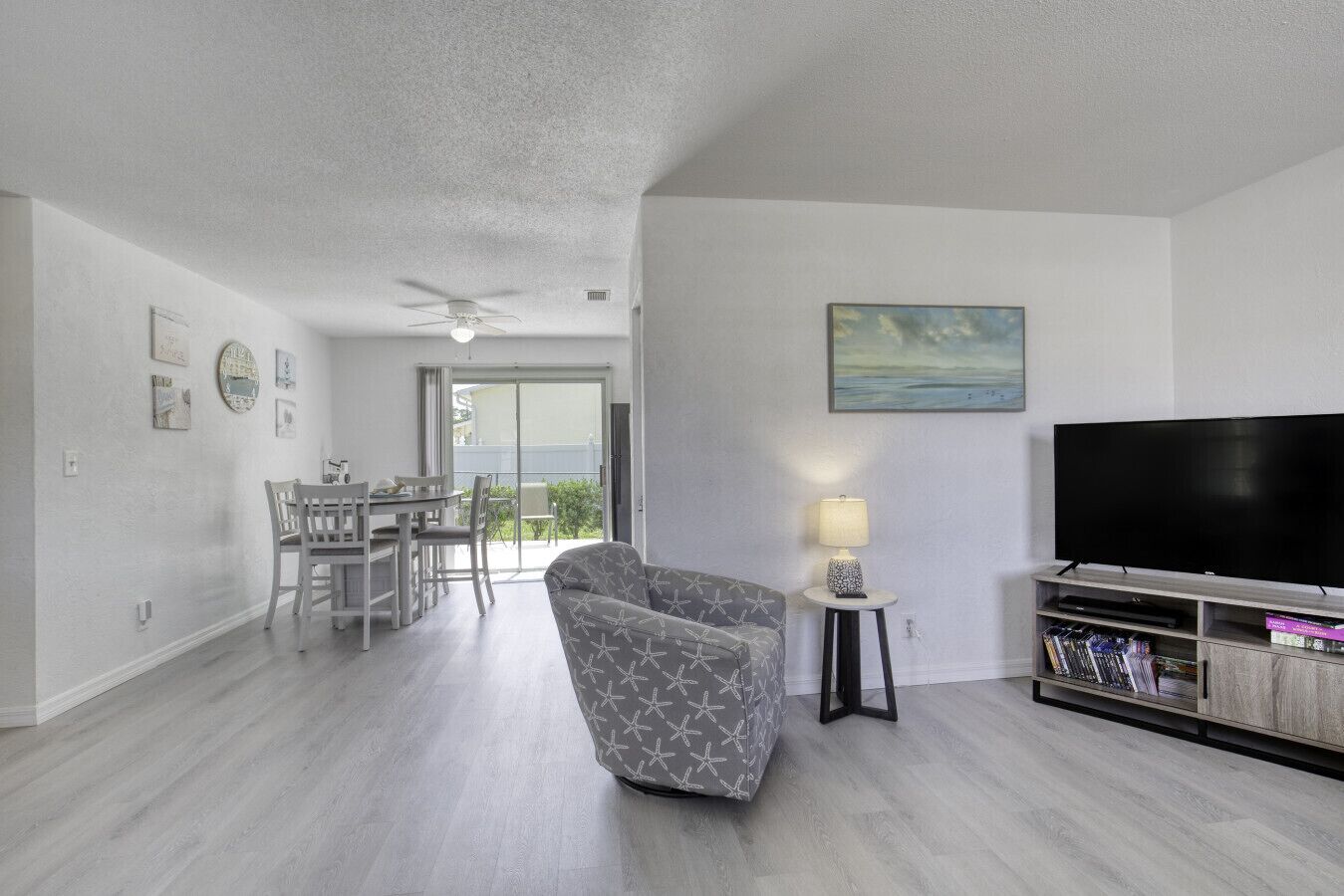 A modern living room with a flat-screen TV, a patterned armchair, a dining area with a table and chairs, and a balcony with a view.