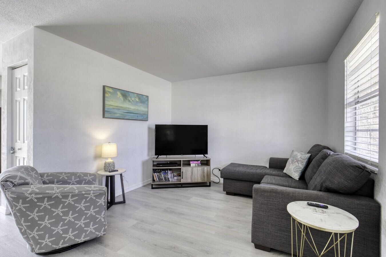 Un salon avec un canapé gris, un fauteuil à motifs, un téléviseur et une petite table avec une lampe.