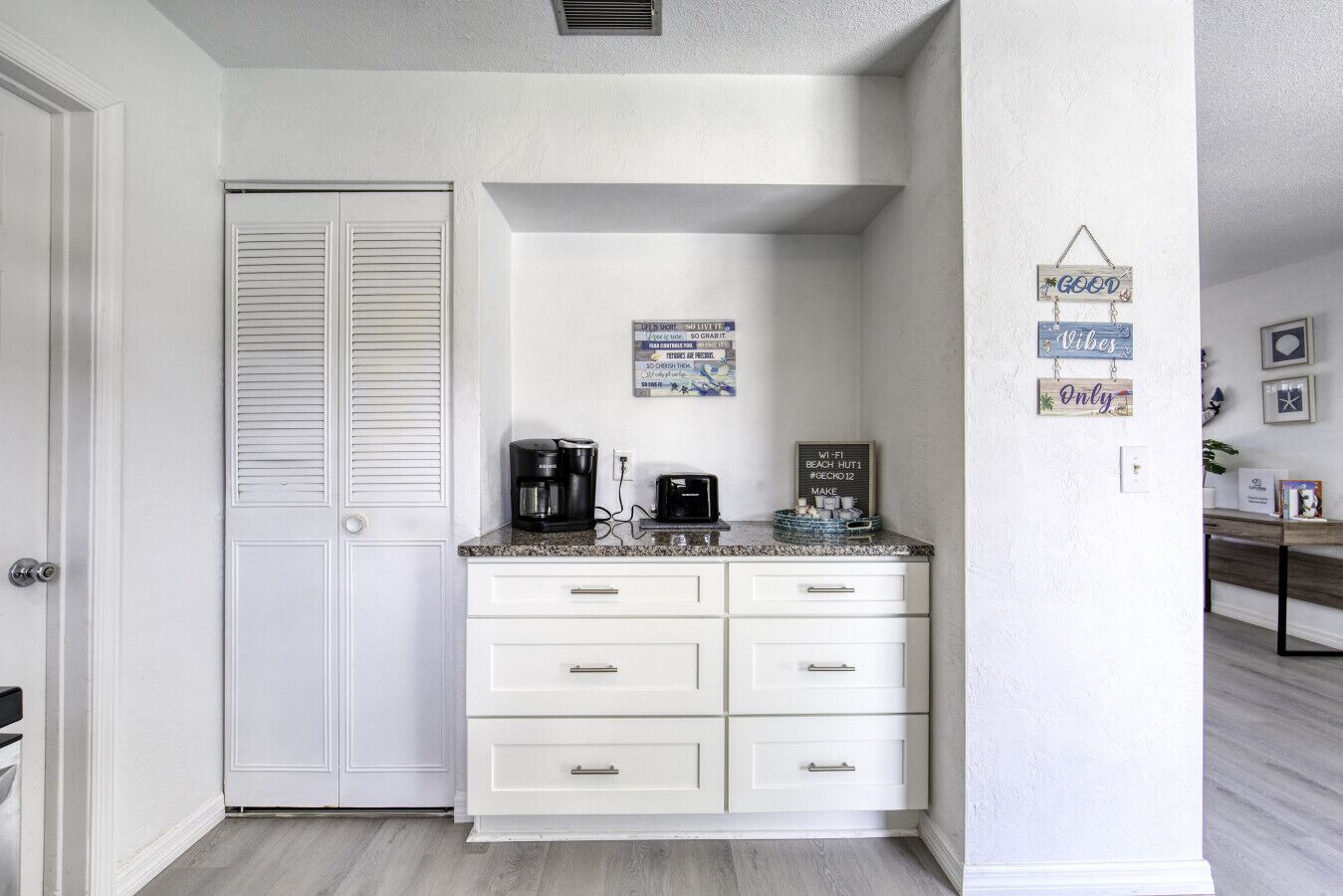 Cocina con armarios blancos, cafetera y tostadora. Hay decoraciones en la pared y un cartel con información de Wi-Fi.