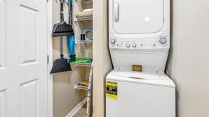 A laundry room with a front-loading washing machine, a broom, a dustpan, and a shelf with cleaning supplies. - Coastside (Santa Rosa Beach)