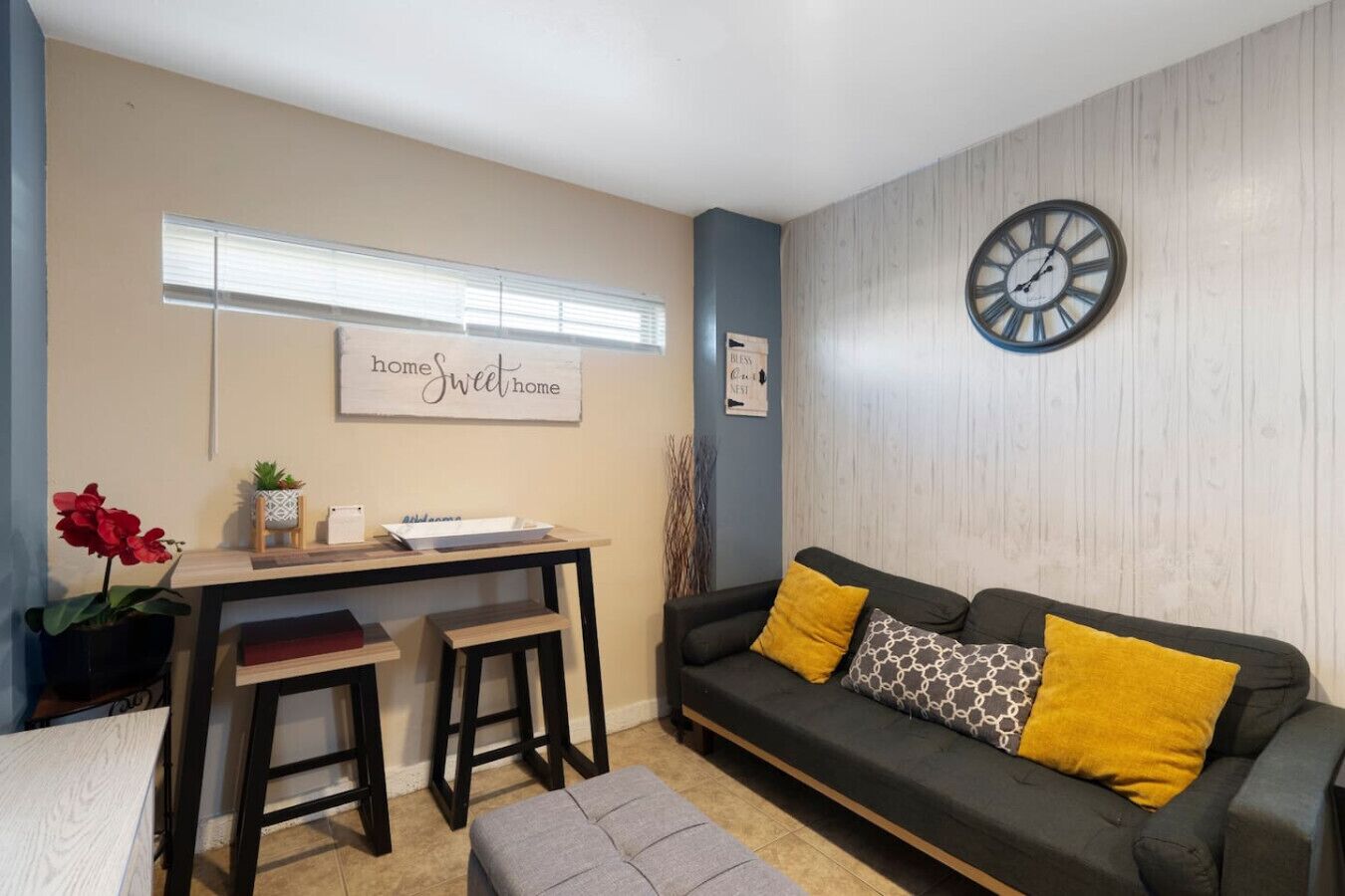 A cozy living room with a dark grey sofa, two yellow cushions, a small wooden table with a plant, and a wall clock.