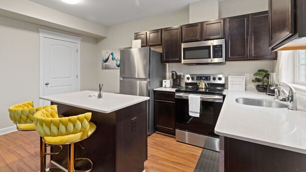 A modern kitchen with dark wooden cabinets, stainless steel appliances, and a white countertop. There are two yellow bar stools and a white door in the background. - Cozy and Comfortable 1-br in the Heart of Detroit (Detroit)