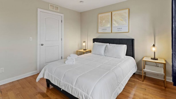 A neatly arranged bedroom with a large bed, two bedside tables with lamps, a door, and two framed maps on the wall. - Cozy and Comfortable 1-br in the Heart of Detroit (Detroit)