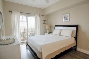 A bedroom with a large bed, a bedside table, a lamp, a window with curtains, and a chair by the window. - Coconut Grove - 403 (North Myrtle Beach)