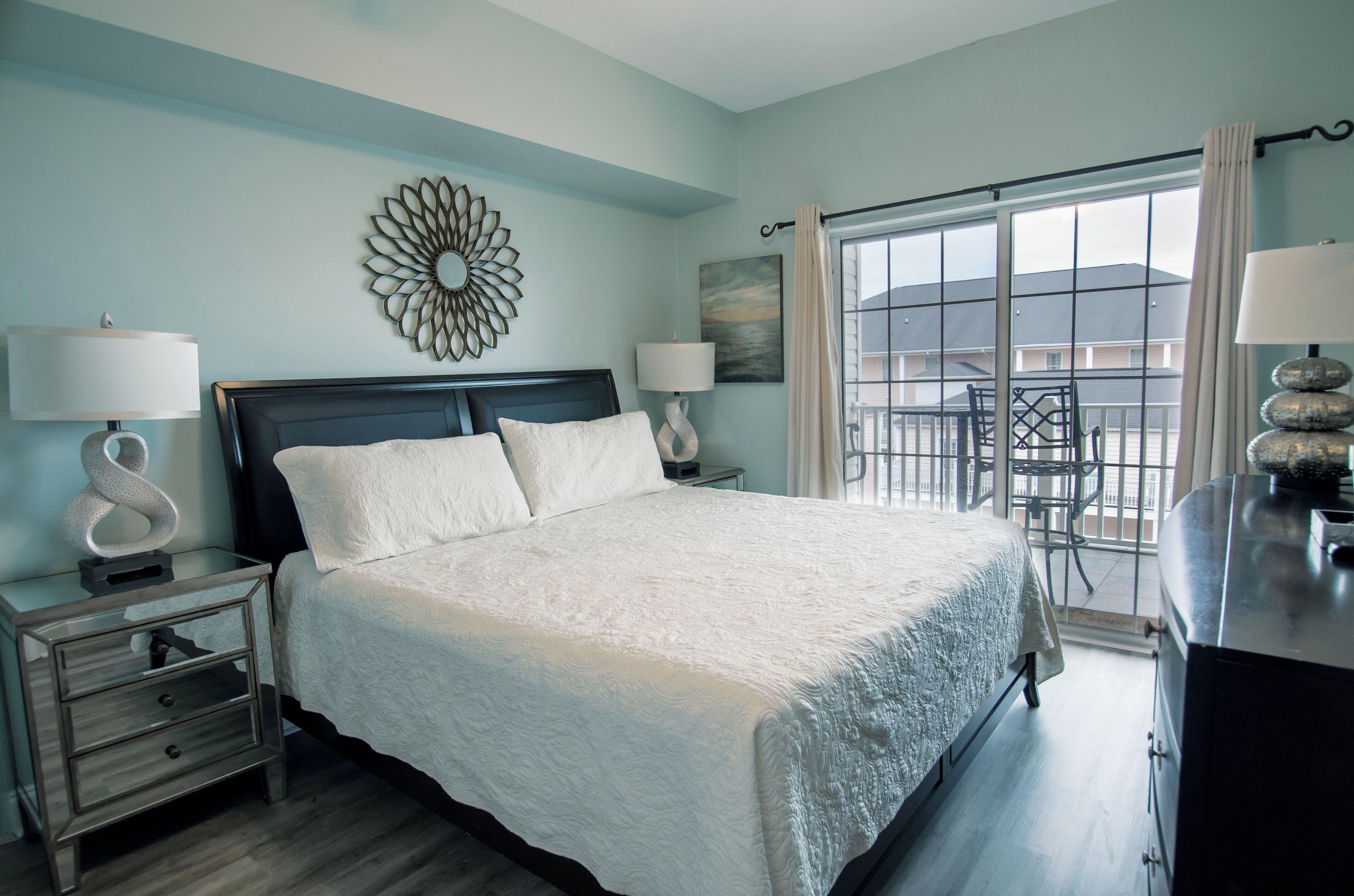 A bedroom with a large bed, two bedside tables with lamps, a decorative mirror, and a view of the outside through a large window.