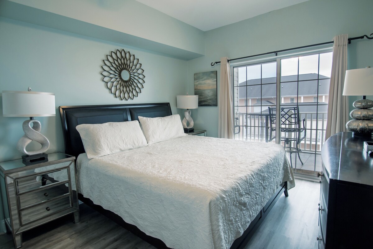 A bedroom with a large bed, two bedside tables with lamps, a decorative mirror, and a view of the outside through a large window.
