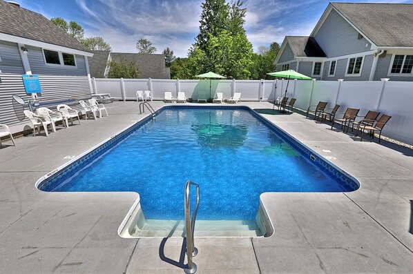 Una piscina rectangular con una escalera metálica, rodeada de sillas reclinables blancas y marrones, y una valla blanca.