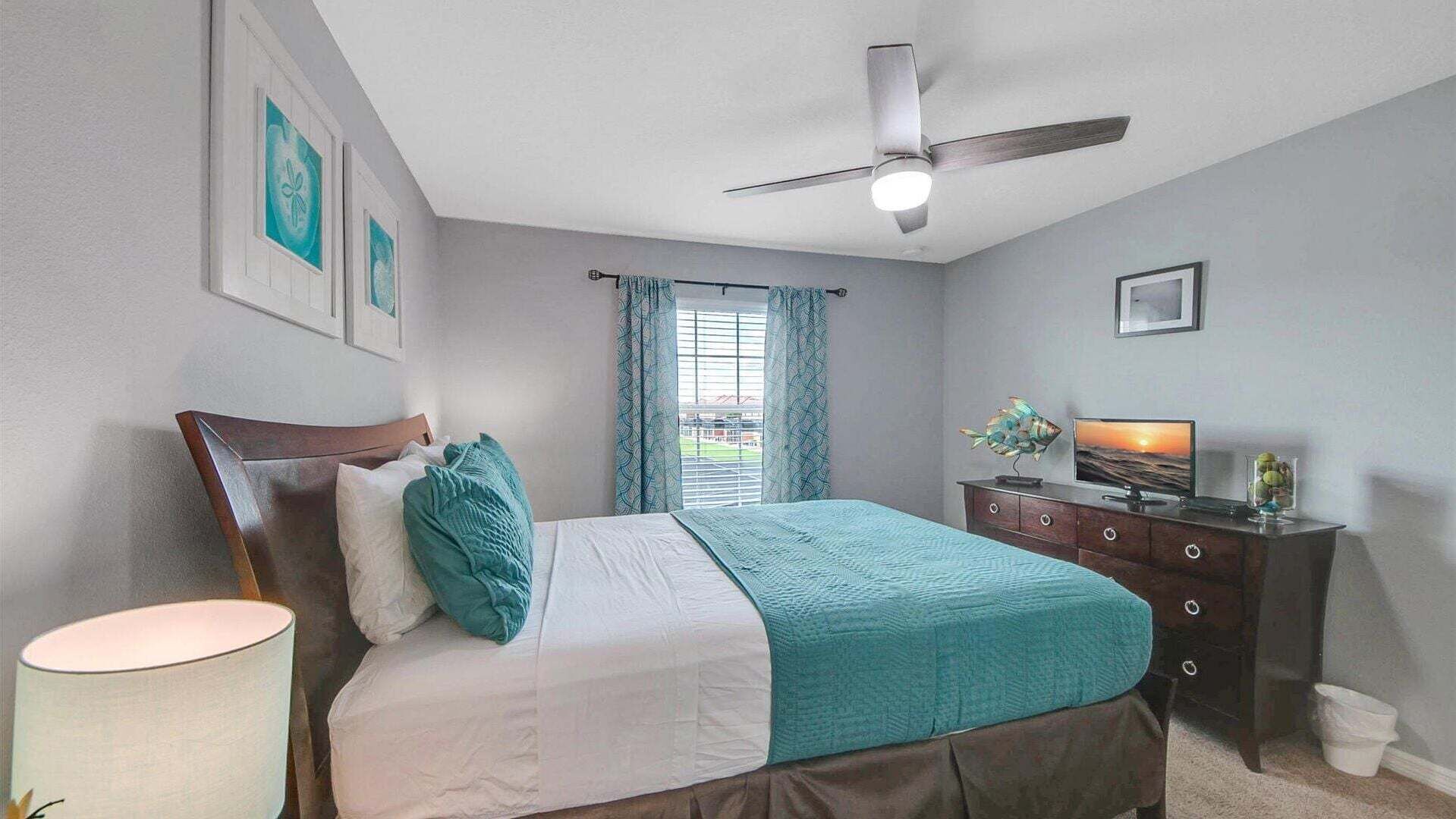 Une chambre à coucher avec un lit, un ventilateur de plafond, une commode, un téléviseur et une fenêtre avec des rideaux.