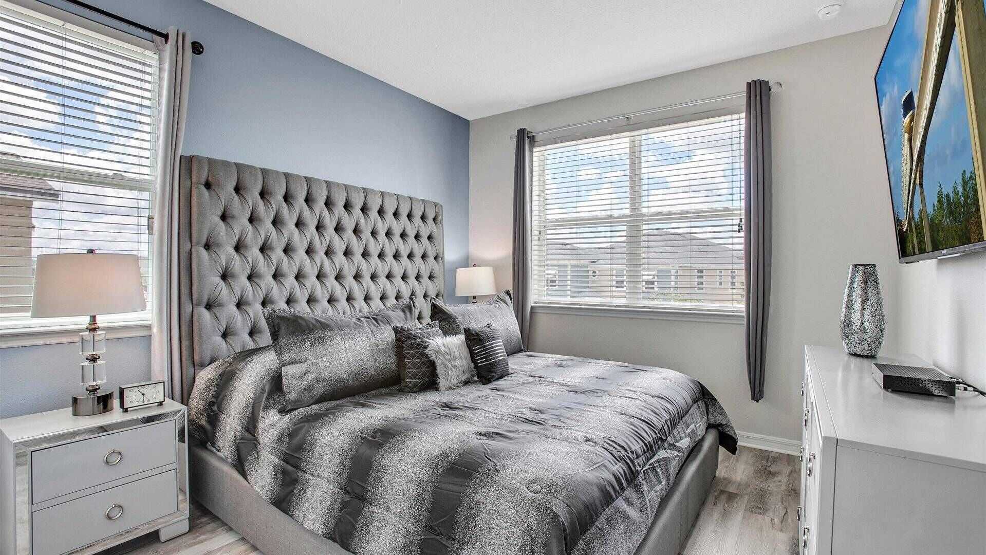 A bedroom with a tufted headboard, a bed with a patterned bedspread, bedside tables with lamps, and a television on the wall.