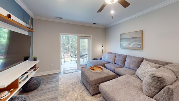 A modern living room with a sectional sofa, a flat-screen TV, a ceiling fan, and a large window with a view of the outdoors. - Crow Creek 1706 (Calabash)