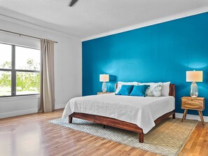 A bedroom with a large bed, two bedside lamps, a window with curtains, and a view of greenery outside. - Coastal Oasis 3 (Nokomis)