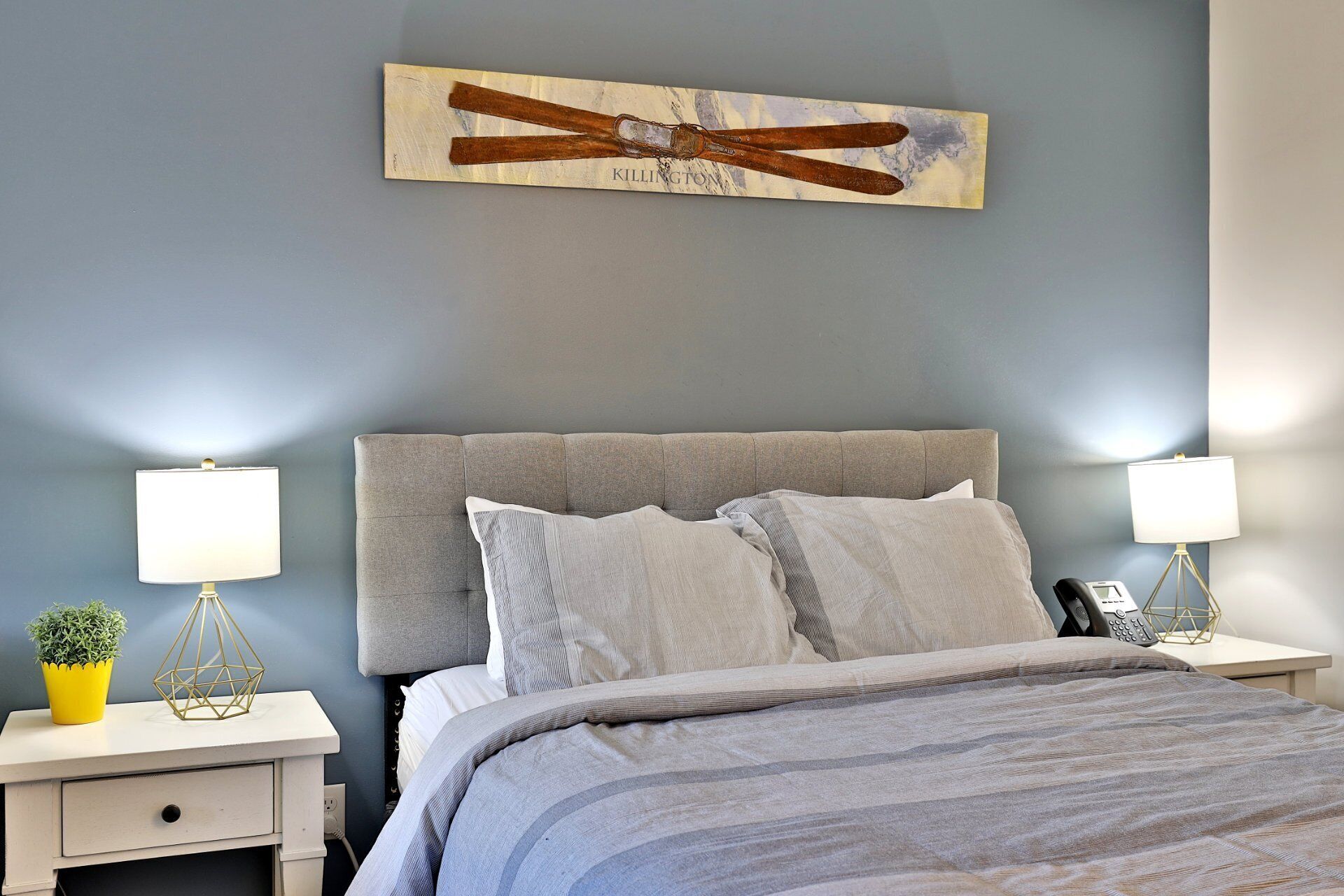Un dormitorio con cabecera gris, dos mesitas de noche con lámparas, cama con ropa de cama gris y una obra de arte colgada en la pared.