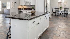 Une cuisine spacieuse avec des meubles blancs, un plan de travail en granit et un coin repas agrémenté d’un lustre.