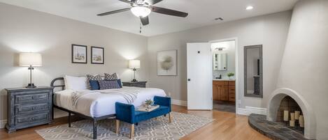A bedroom with a bed, bedside tables, a ceiling fan, a fireplace, and a hallway leading to a bathroom.