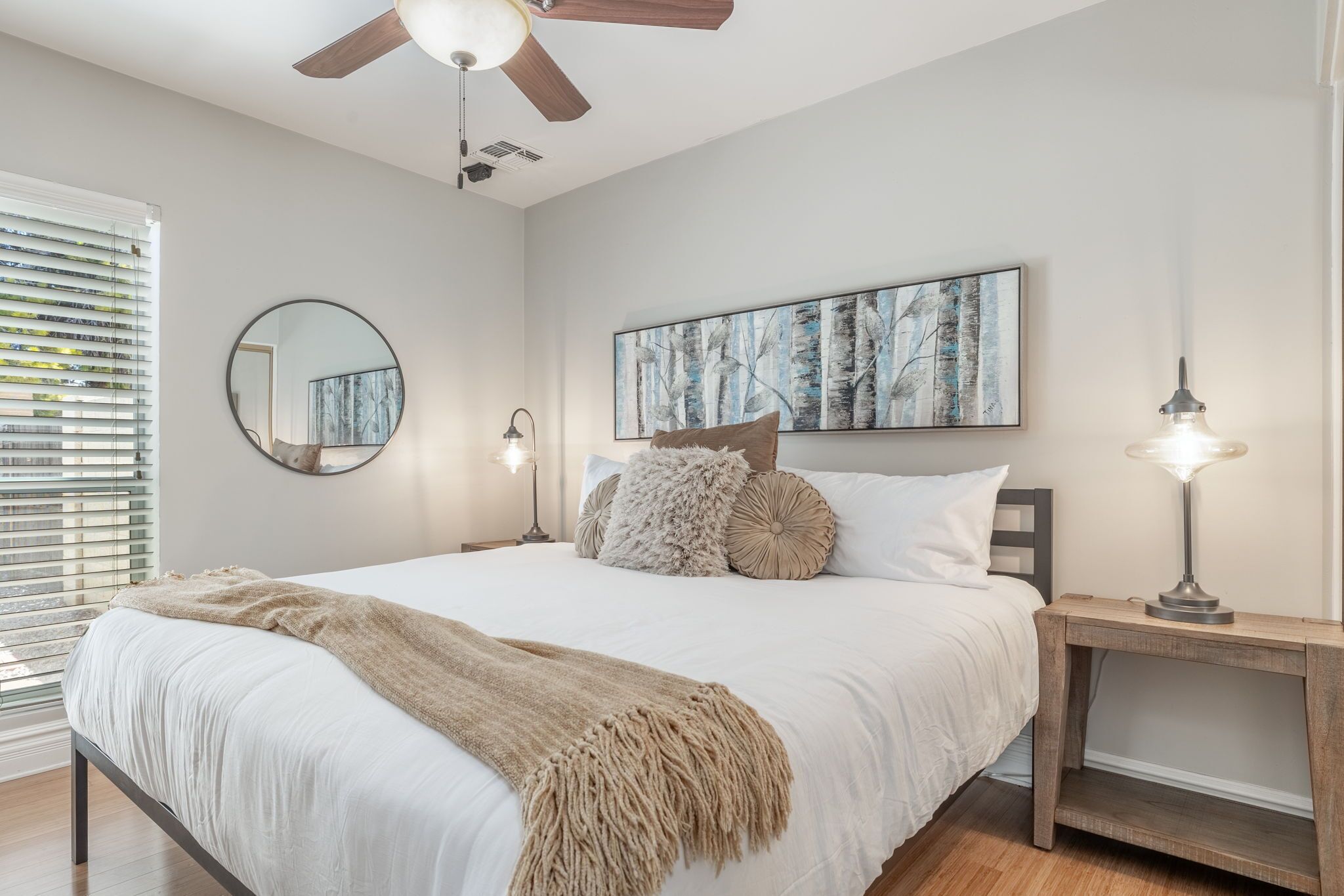 Une chambre à coucher comprenant un lit, un miroir rond, un ventilateur de plafond, une table de chevet avec une lampe et un tableau accroché au mur.