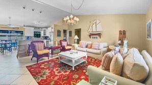 Ein geräumiger Wohnbereich mit einem großen, roten Teppich mit Blumenmuster, einem weißen Couchtisch und einem beigefarbenen Sofa. Im Hintergrund ist eine Küche mit modernen Geräten und ein Essbereich mit einer Bar zu sehen.