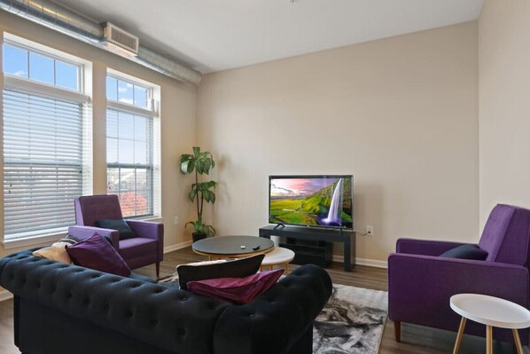 A living room with a black sofa, two purple armchairs, a round coffee table, and a TV displaying a scenic landscape. - Cozy 3bd, 3BA Downtown Indy (Indianapolis)
