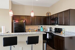 Una cocina moderna con armarios de madera oscura, un refrigerador negro, un microondas y una encimera de granito. Hay dos taburetes negros y una lámpara colgante.