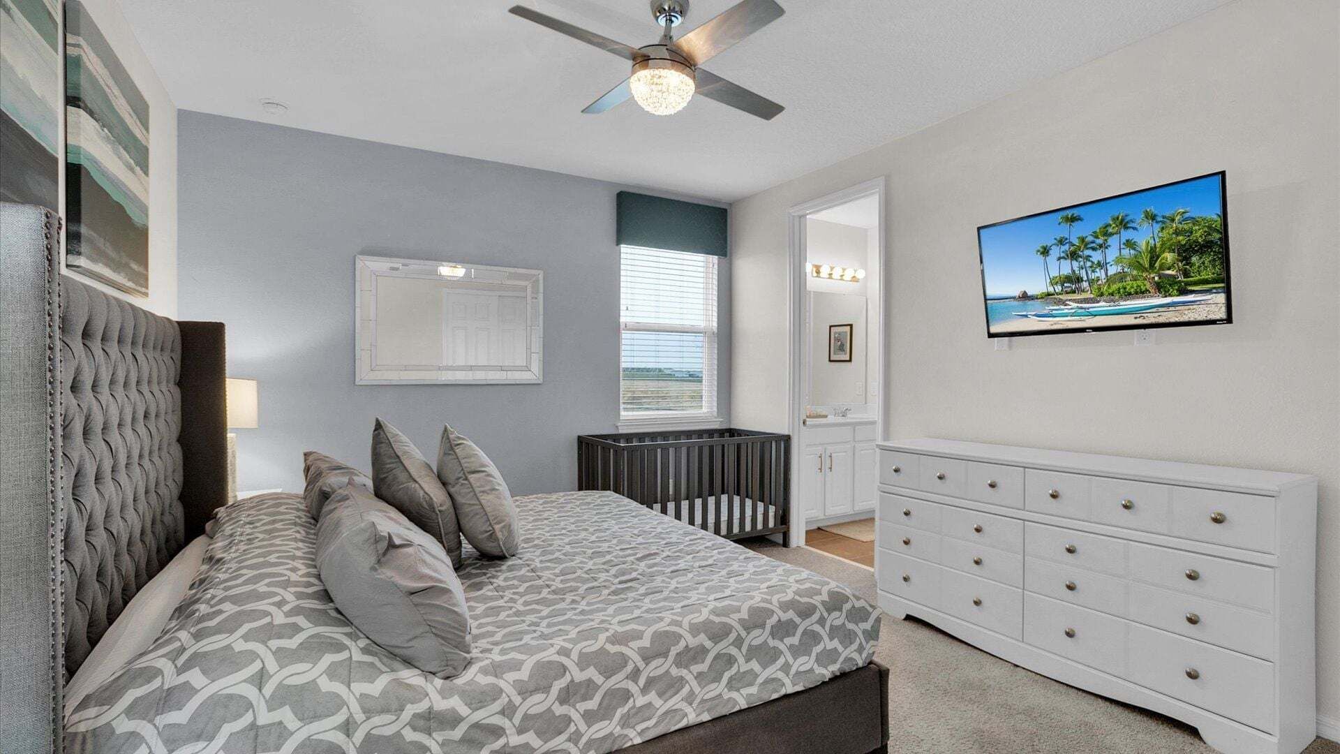 Un dormitorio con cama, cuna, cómoda, ventilador de techo y televisor que muestra una escena de playa.