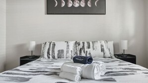 A hotel room with a bed featuring a black and white abstract pattern, two nightstands with lamps, a wall-mounted painting of the moon phases, and neatly folded towels on the bed. - Cozy 3BR Home Family-friendly Near Attractions (San Antonio)