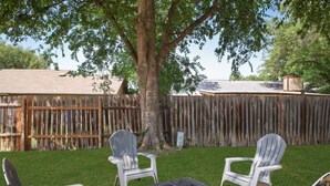 A backyard with a wooden fence, a large tree, and four Adirondack chairs around a small table. - Cozy 3BR Home Family-friendly Near Attractions (San Antonio)