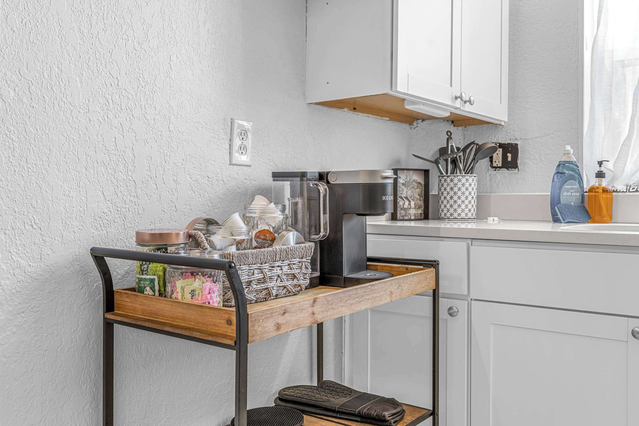A kitchen with white cabinets, a coffee machine, a kettle, and a basket of snacks on a wooden shelf.