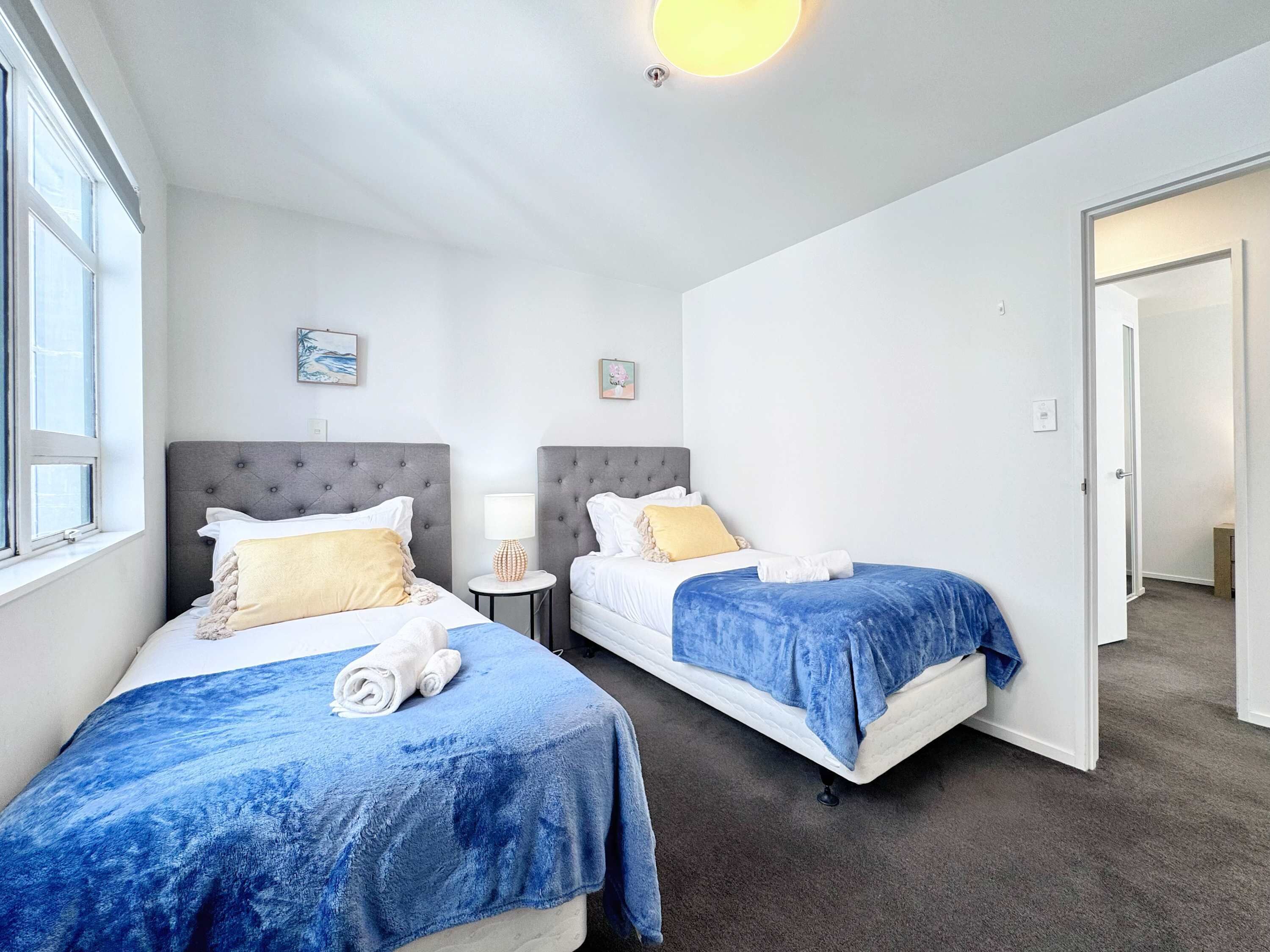 Une chambre d’hôtel avec deux lits, chacun recouvert d’une couverture bleue et de draps blancs. Une petite table avec une lampe se trouve entre les lits, et un tableau est accroché au mur.