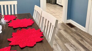 Un comedor con una mesa de madera, manteles individuales rojos y sillas blancas. Se ve una cocina a través de un vano abierto.
