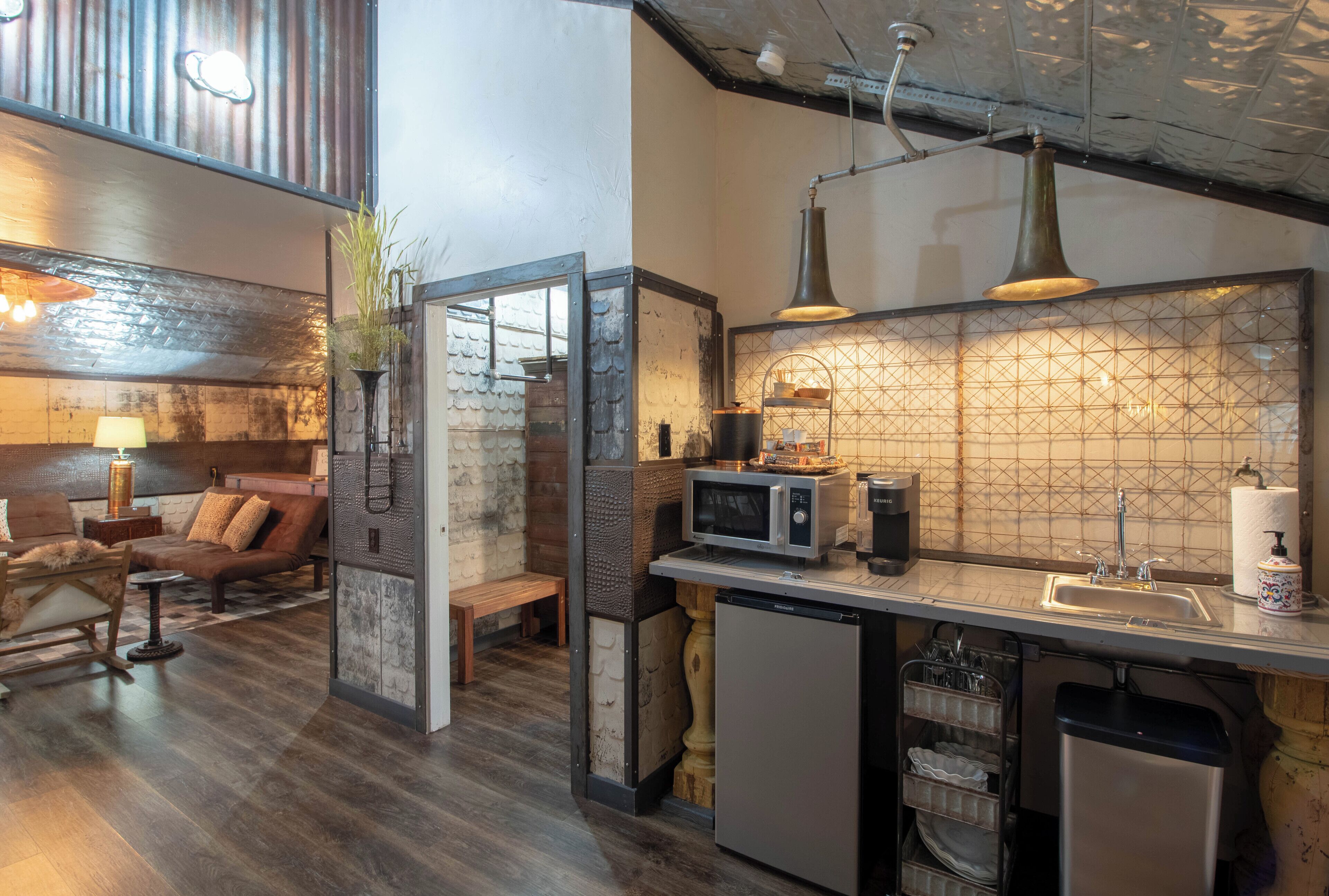 Cocina compacta con microondas, lavabo y escurreplatos. Una pequeña zona de estar con sofá y lámpara se encuentra adyacente a la cocina.