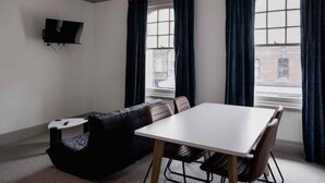 Une pièce avec une table blanche et quatre chaises, un canapé noir, un téléviseur à écran plat fixé au mur et de grandes fenêtres avec des rideaux foncés.