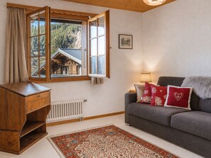 A cozy living room with a wooden bookshelf, a grey sofa with red cushions, a patterned rug, and a window with a view of a mountain. - The Aigle Royal residence is located next to the Barboleuse gondola which takes you to the Gryon - V (Gryon)